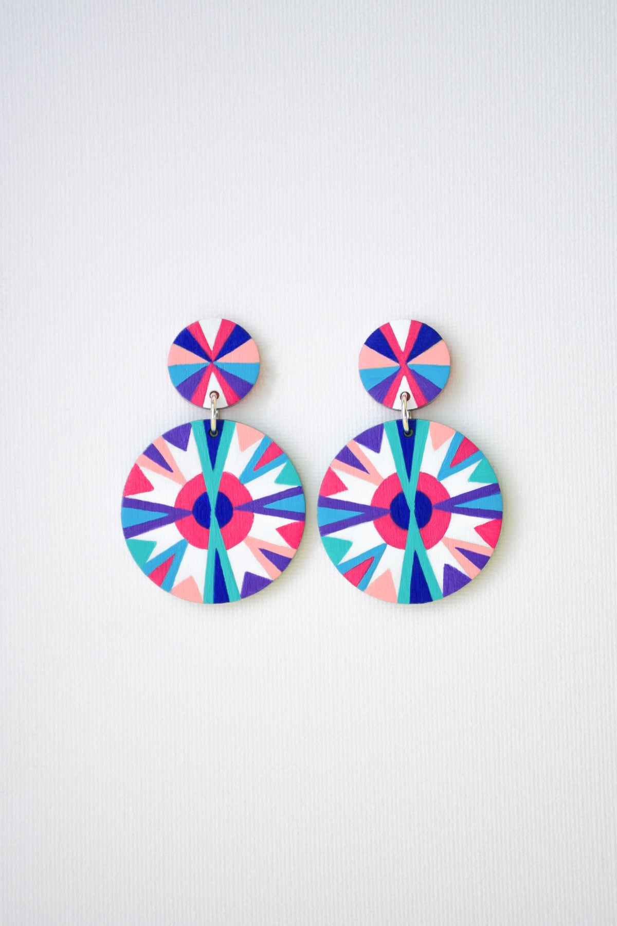 A pair of drop earrings with colorful geometric starburst patterns in pink, blue, purple, and white, displayed against a light gray textured background.