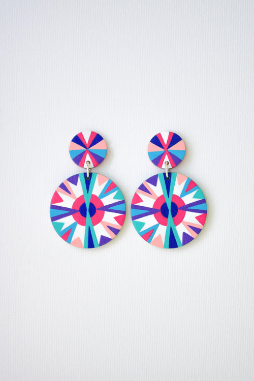 A pair of drop earrings with colorful geometric starburst patterns in pink, blue, purple, and white, displayed against a light gray textured background.