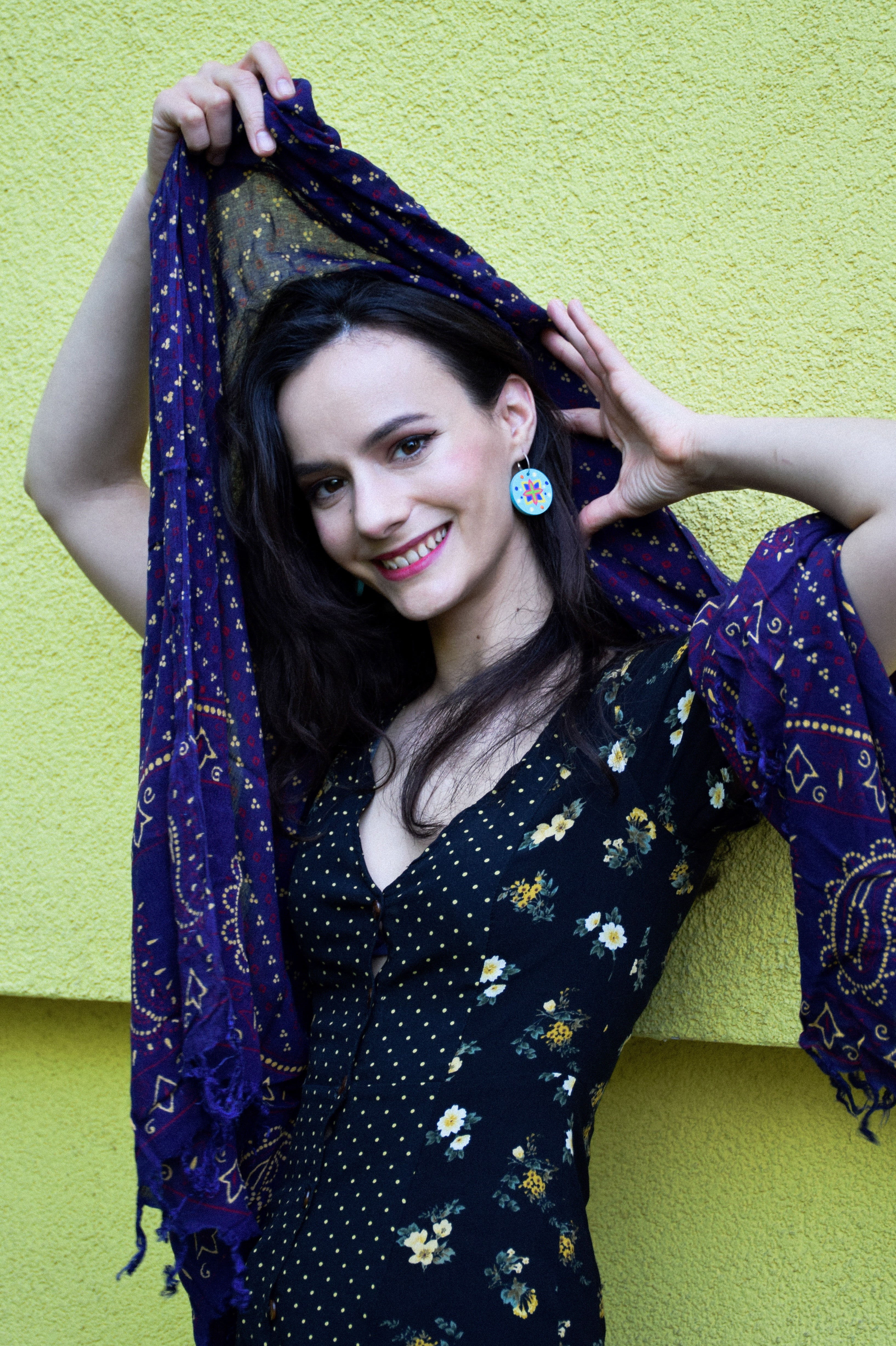 Woman holding a purple scarf and wearing colorful earrings against a yellow wall