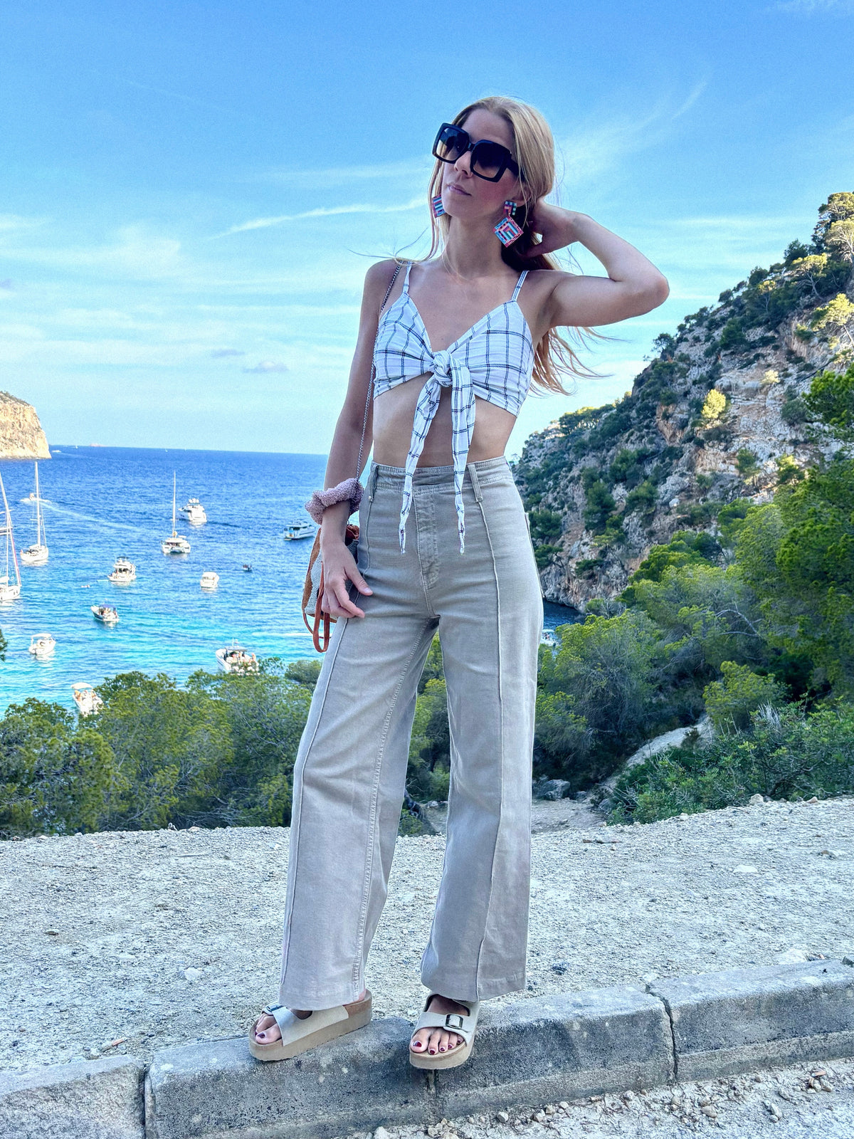 A woman posing outdoors on a stone ledge overlooking a scenic blue sea and green cliffs, wearing a white and black checkered tie-front crop top, beige wide-leg trousers, colorful statement earrings, and beige platform sandals. 