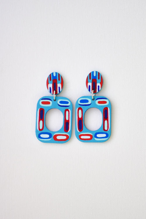A pair of colorful geometric drop earrings with light blue rectangular shapes featuring red and white oval accents, and matching patterned oval studs, displayed on a light textured background.