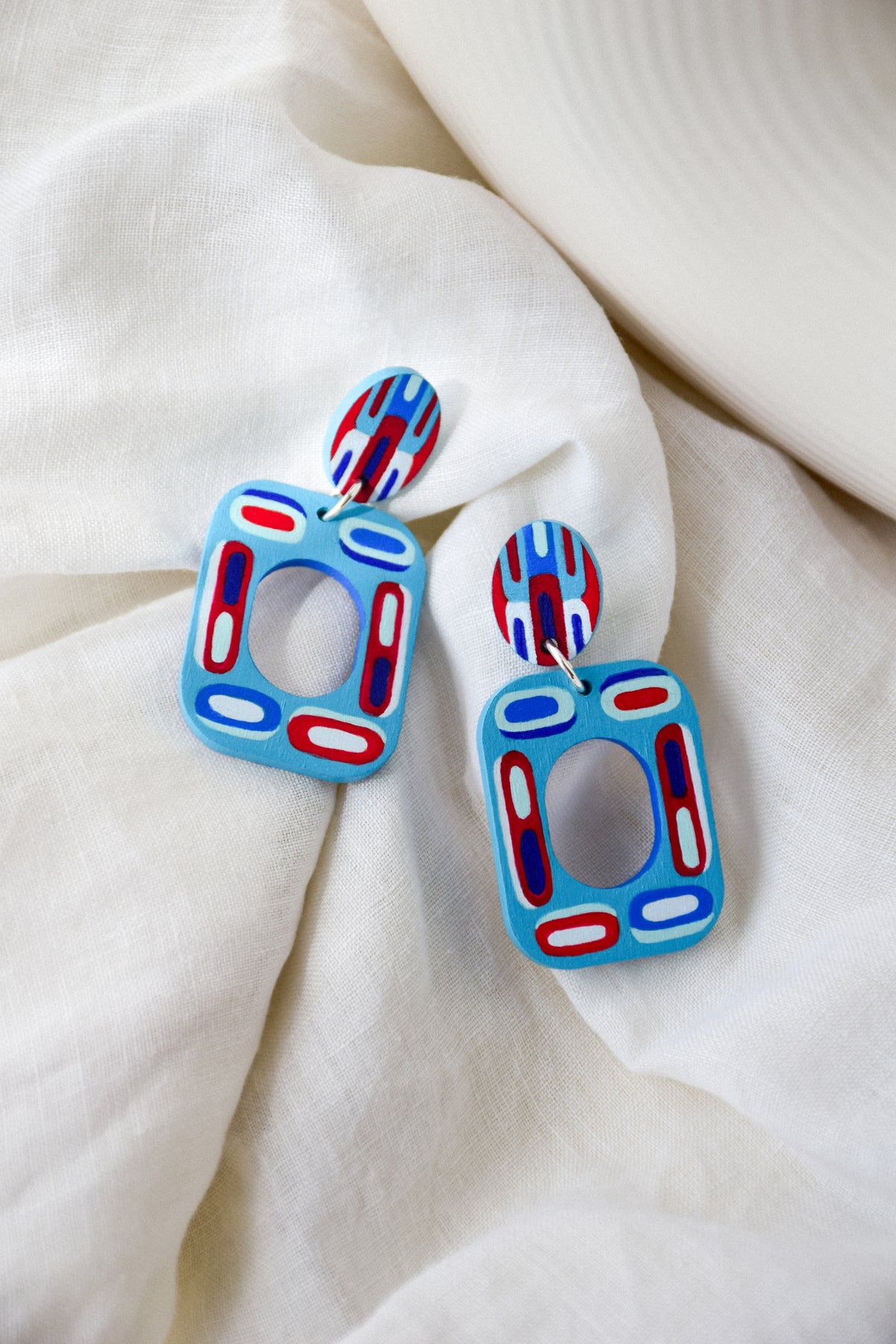 A pair of colorful geometric wooden drop earrings with blue, red, and white patterns, displayed on a soft, draped white fabric background.