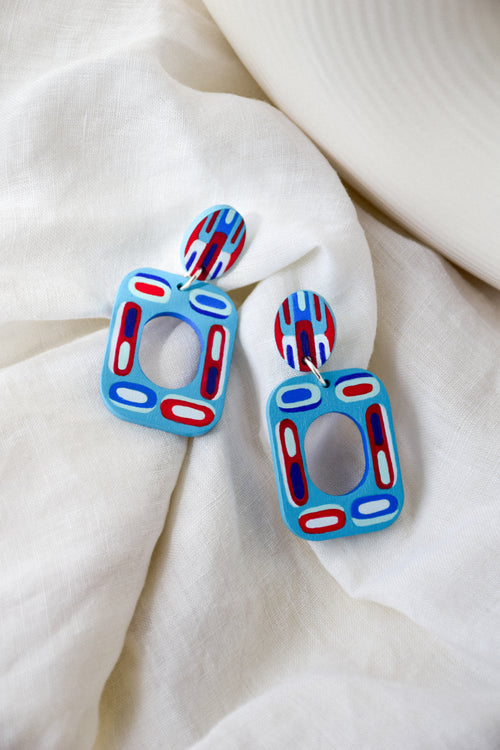 A pair of colorful geometric wooden drop earrings with blue, red, and white patterns, displayed on a soft, draped white fabric background.