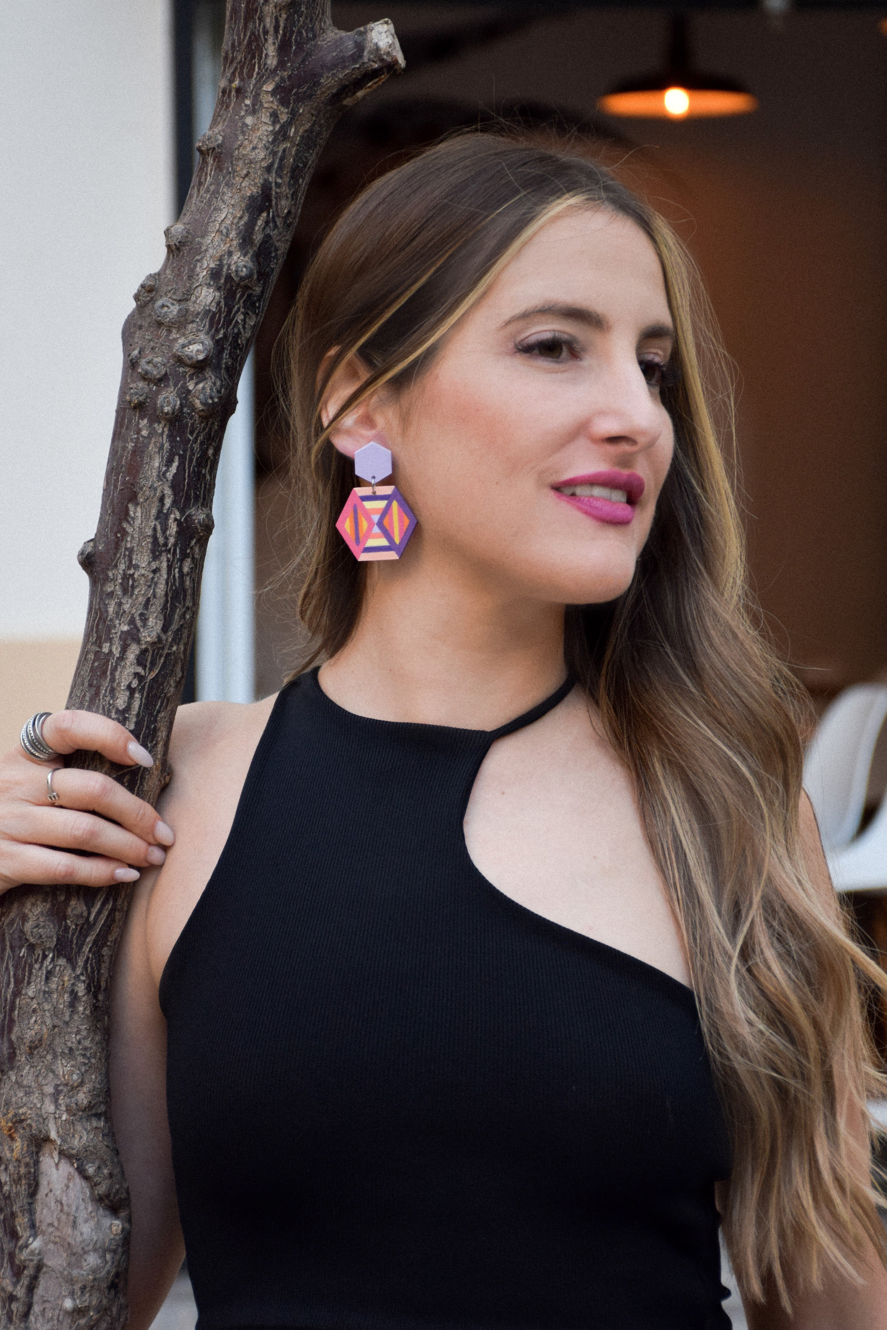 Woman wearing a black top and colorful earrings, standing next to a tree.