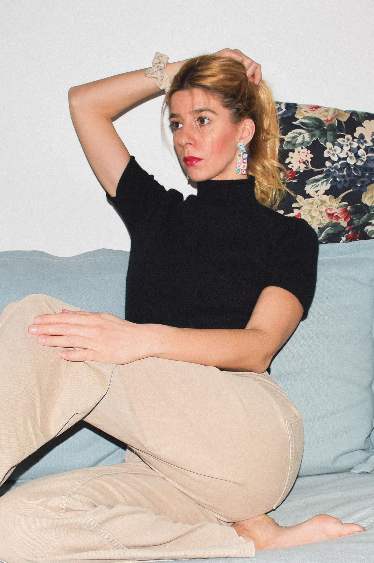 A woman sitting on a light blue couch, wearing a black short-sleeve mock neck top and beige trousers. She has her hair in a ponytail and is wearing colorful dangling earrings.