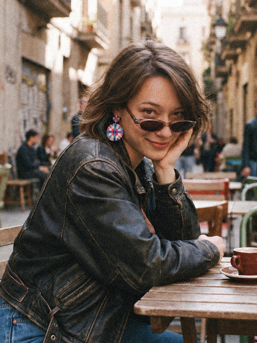 A woman with short brown hair sits at an outdoor cafe table wearing a black leather jacket, dark sunglasses, and colorful geometric earrings. The background shows a blurred street scene with buildings and other cafe patrons.