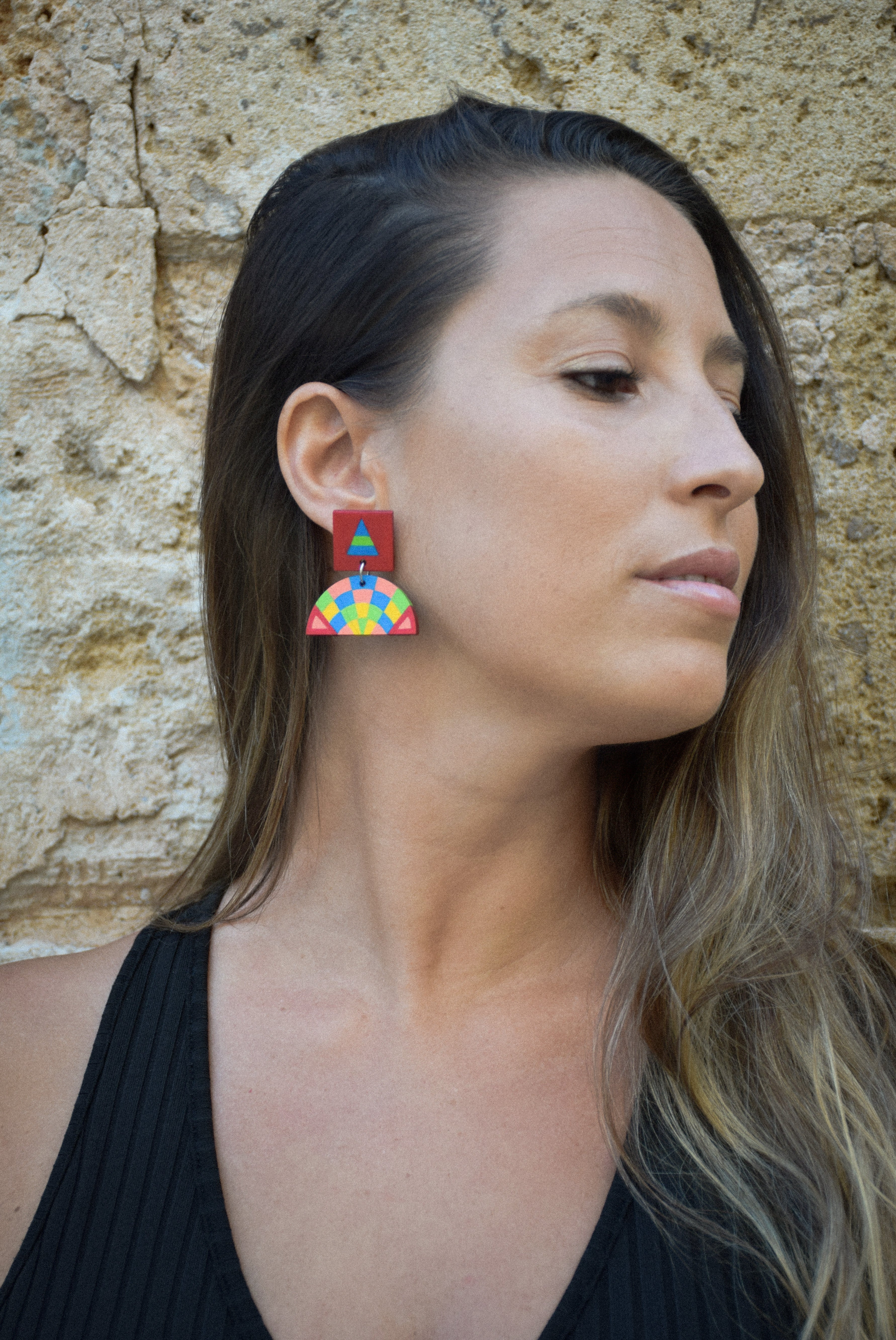 Woman wearing colorful geometric earrings against a stone wall.