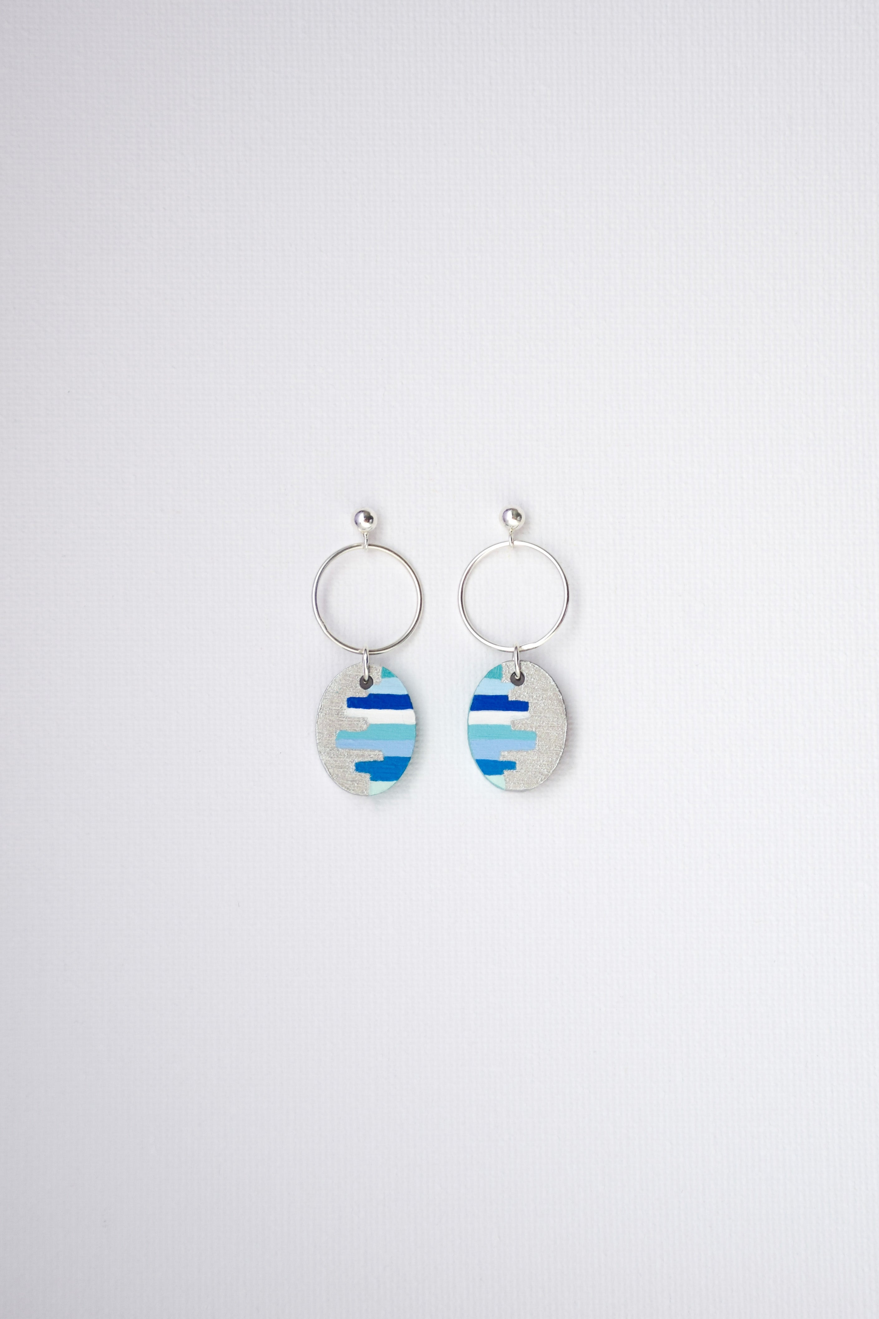 Pair of hand-painted earrings with blue and white striped designs and sterling silver hoops on a light gray background