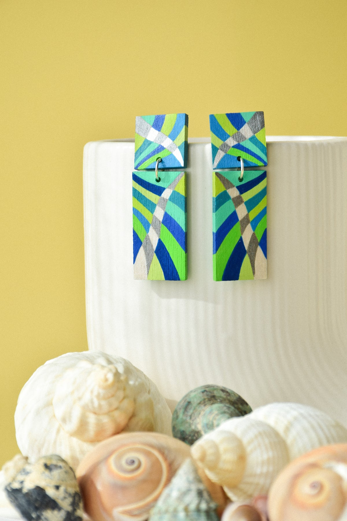 A pair of colorful geometric drop earrings with blue, green, and silver patterns, displayed on a white ribbed vase with seashells in the foreground against a yellow background.