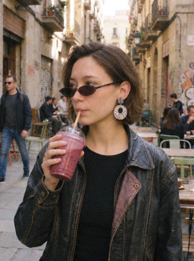 A woman wearing a black leather jacket, black top, round sunglasses, and hand-painted colorful earrings drinks a pink smoothie in a narrow city street with cafe tables and other pedestrians in the background.