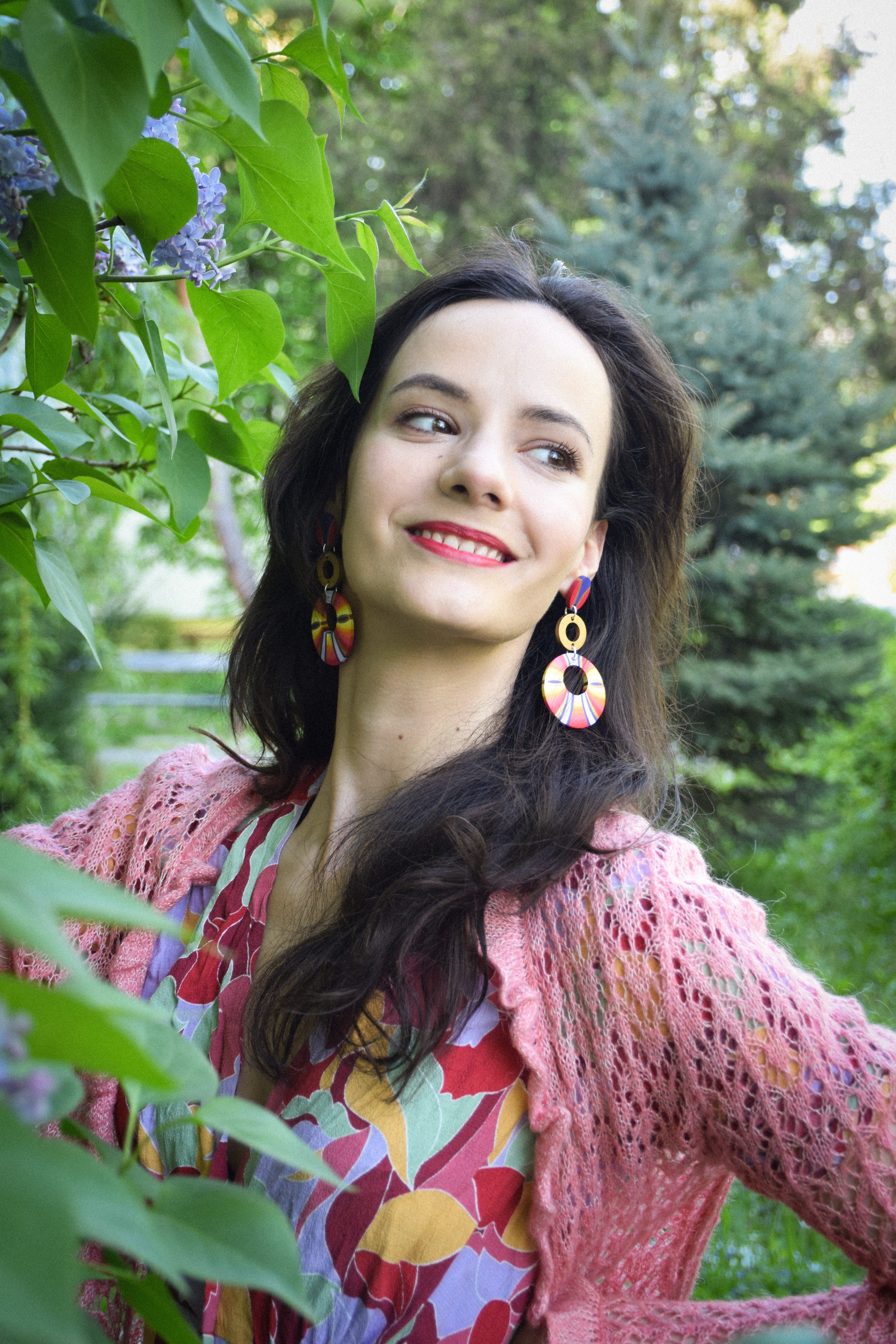 Woman with colorful earrings and a pink cardigan standing among green foliage