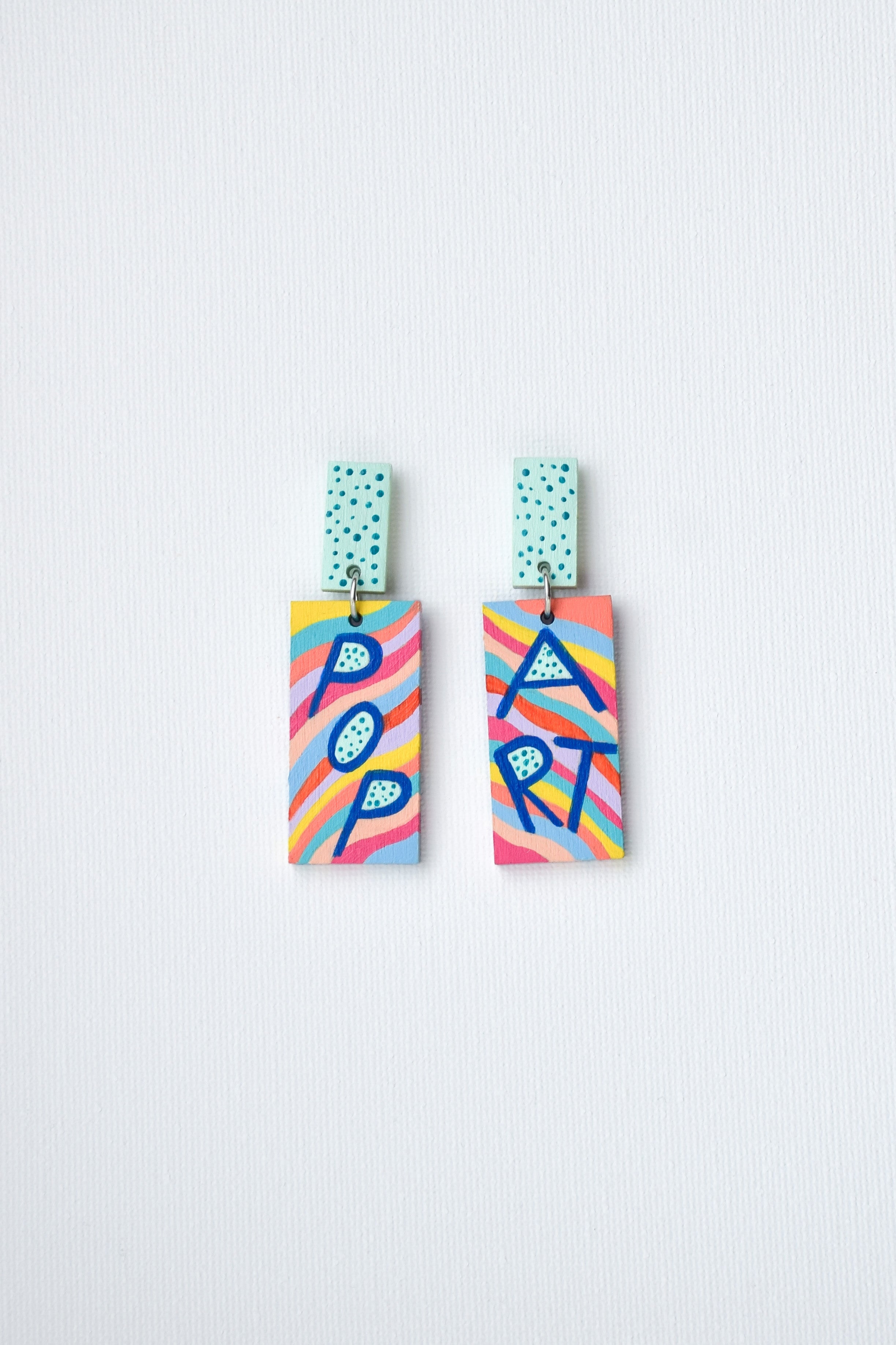 Colorful hand-painted geometric earrings featuring pop art lettering on a light gray background