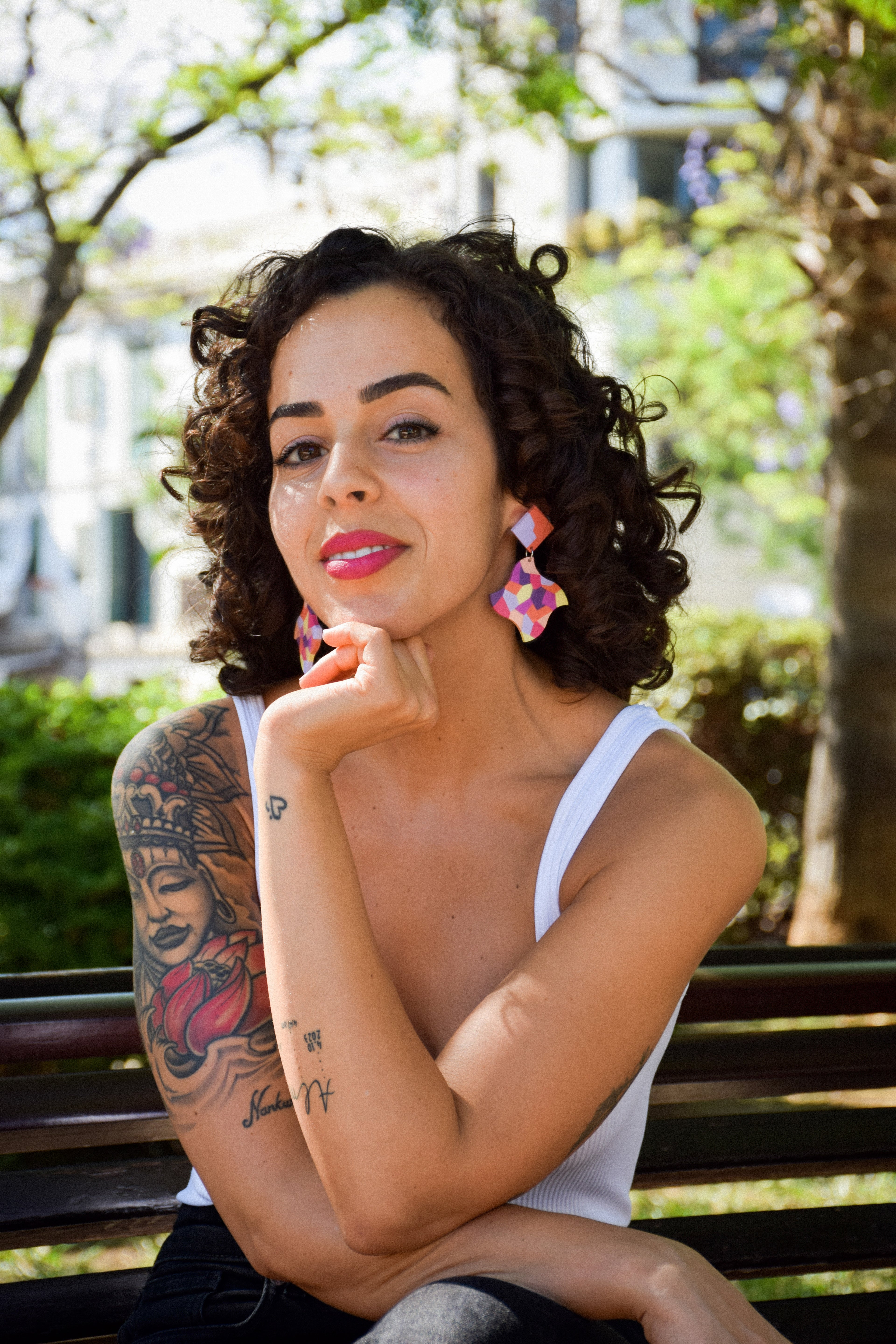 Woman sitting on a bench in an urban park with tattoos and colorful earrings.