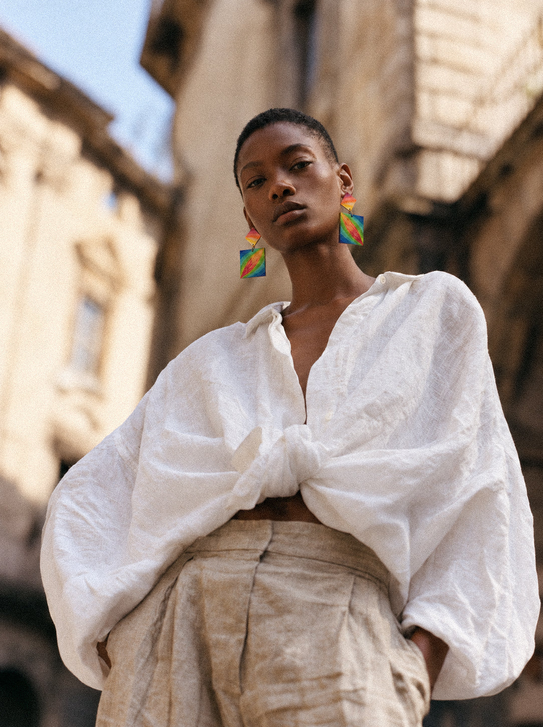A woman wearing oversized white linen shirt, beige linen trousers, and colorful statement earrings in an urban environment. 