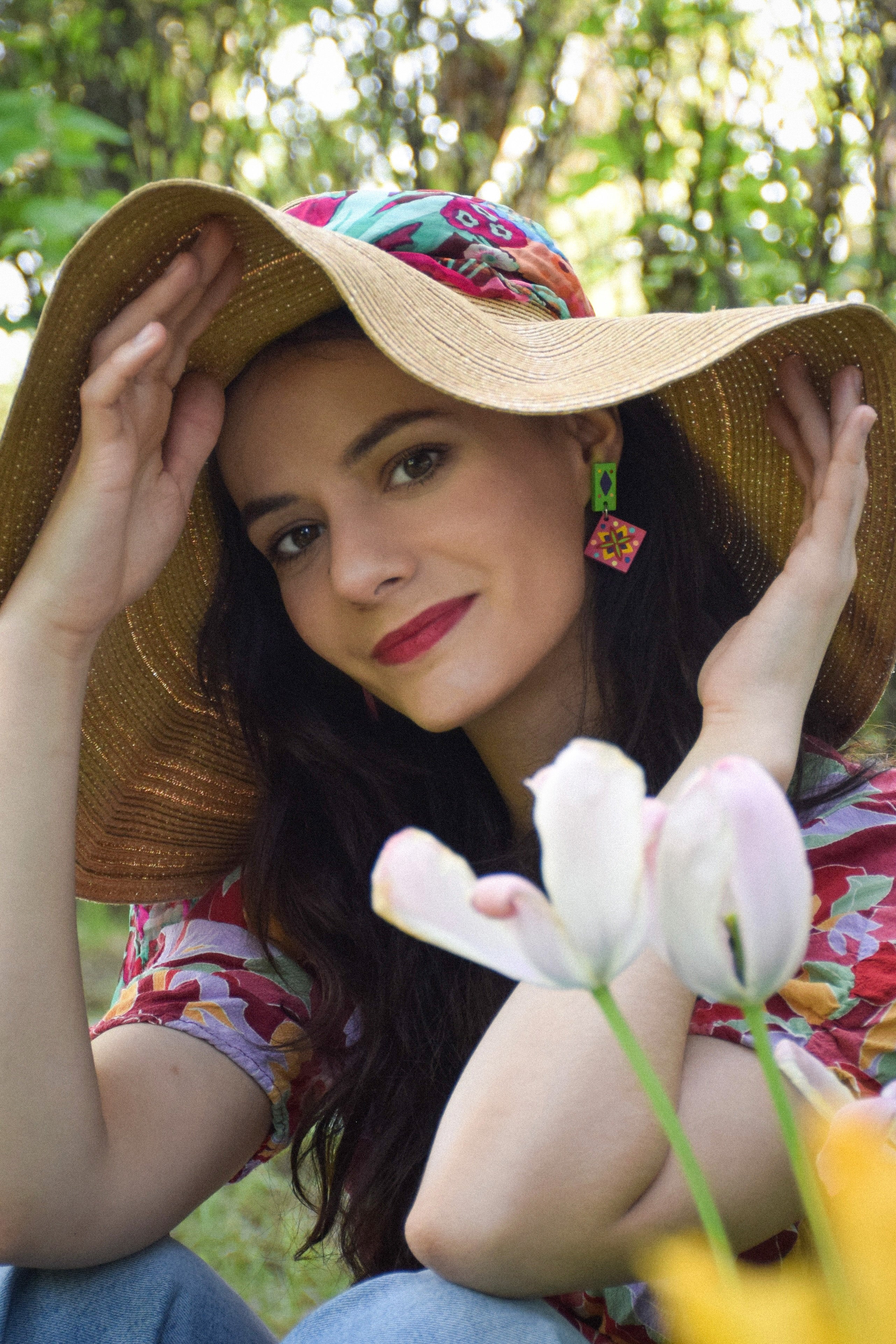 Woman wearing a wide-brimmed hat and colorful earrings, sitting outdoors with flowers.