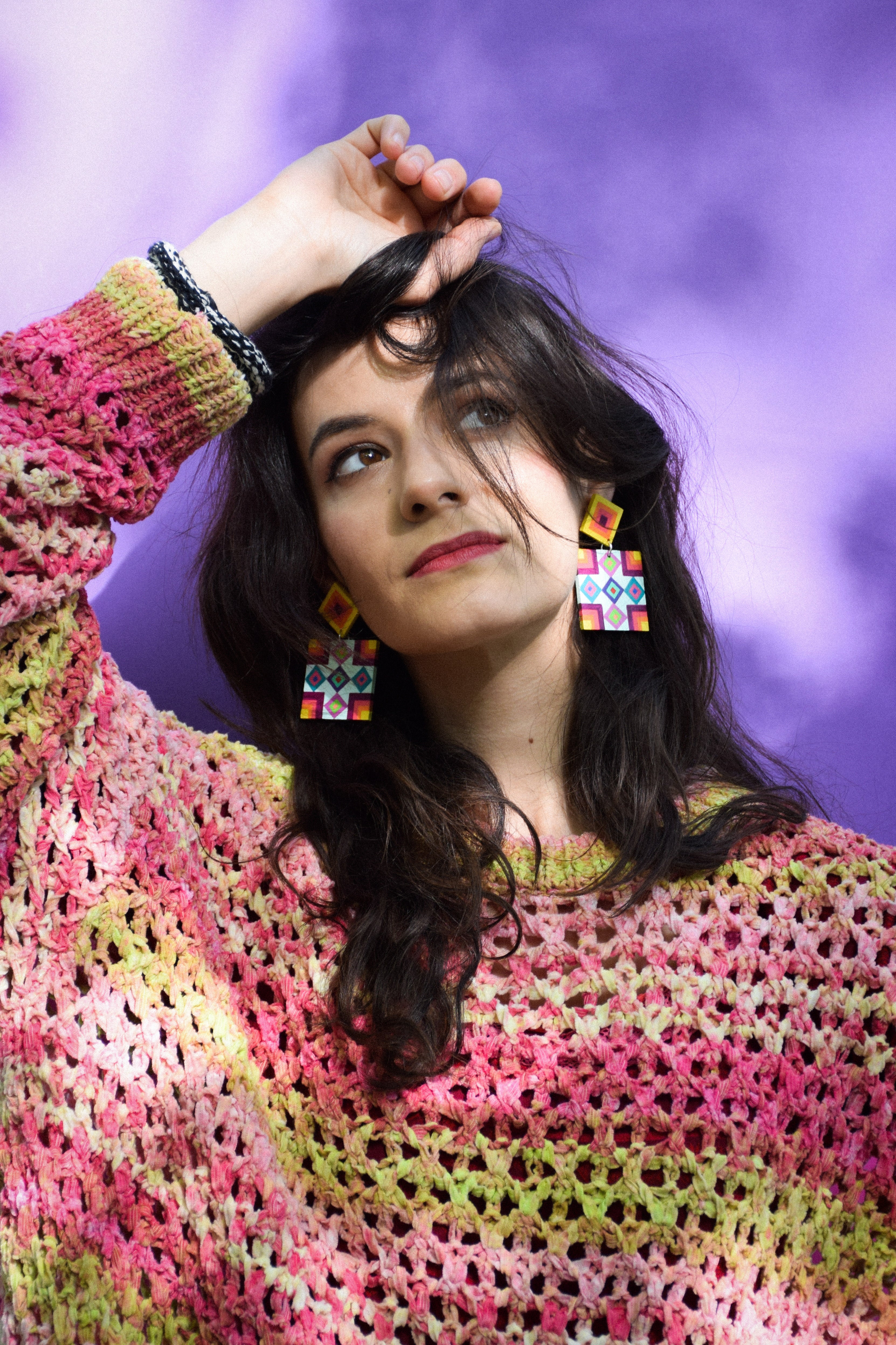 Woman wearing a colorful crochet sweater and colorful statement earrings with a purple background